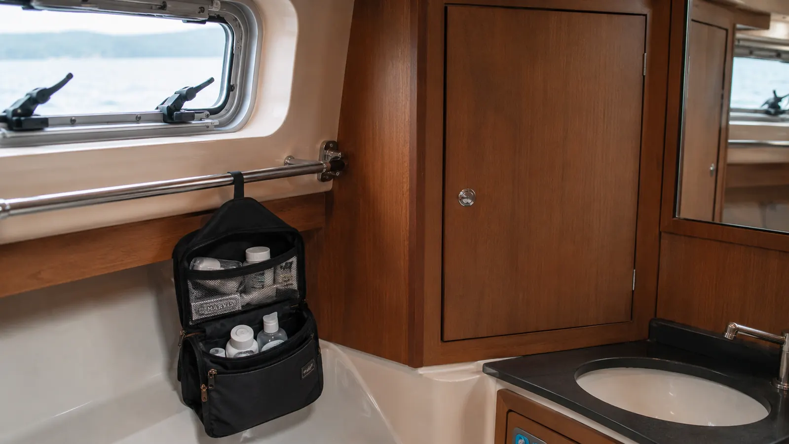 Hanging toiletry bag suspended from a sailboat grab-rail in a compact marine head with port light visible
