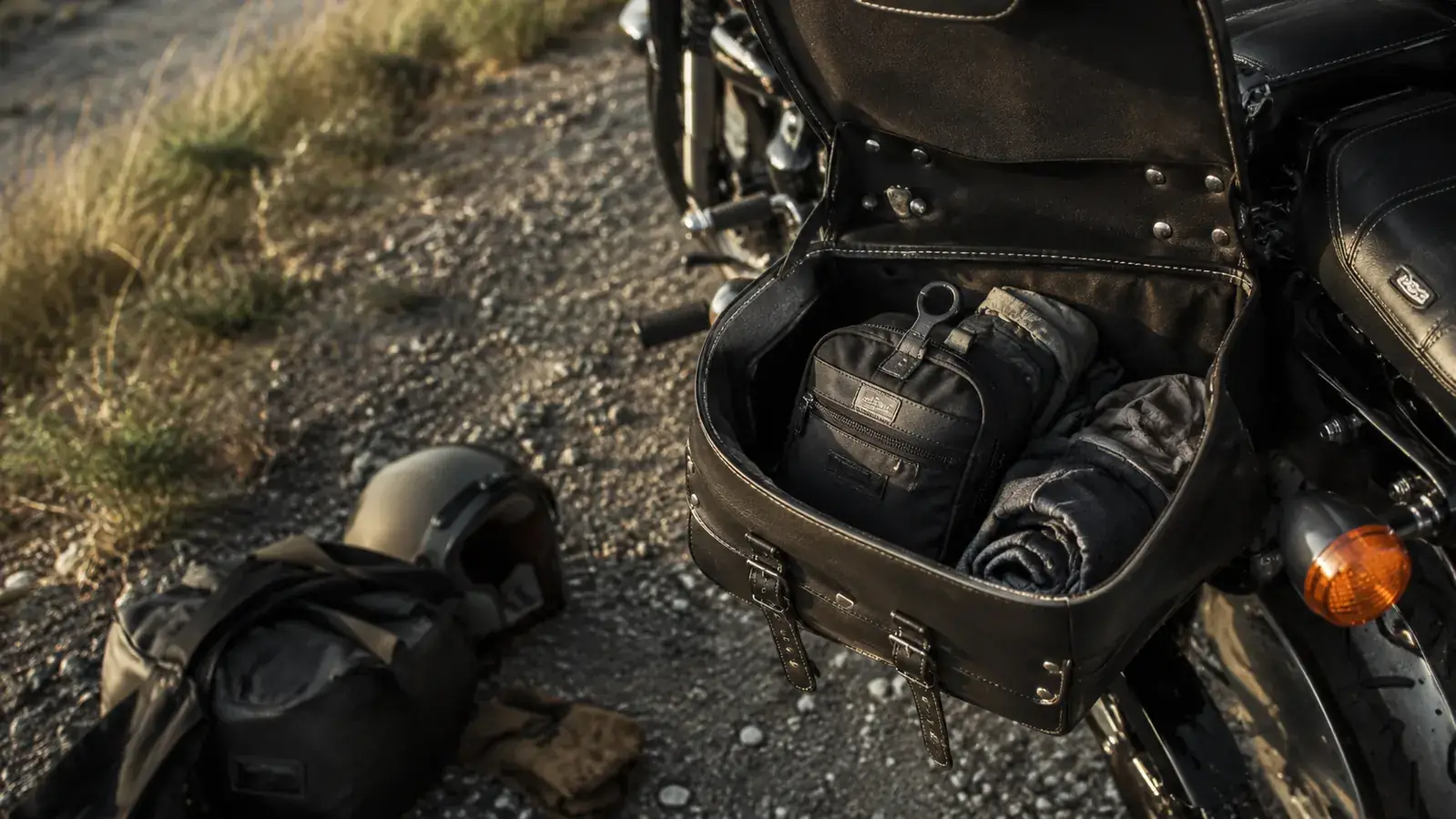 A motorcycle saddlebag open on a roadside, a compact hanging toiletry bag tucked neatly inside alongside road gear